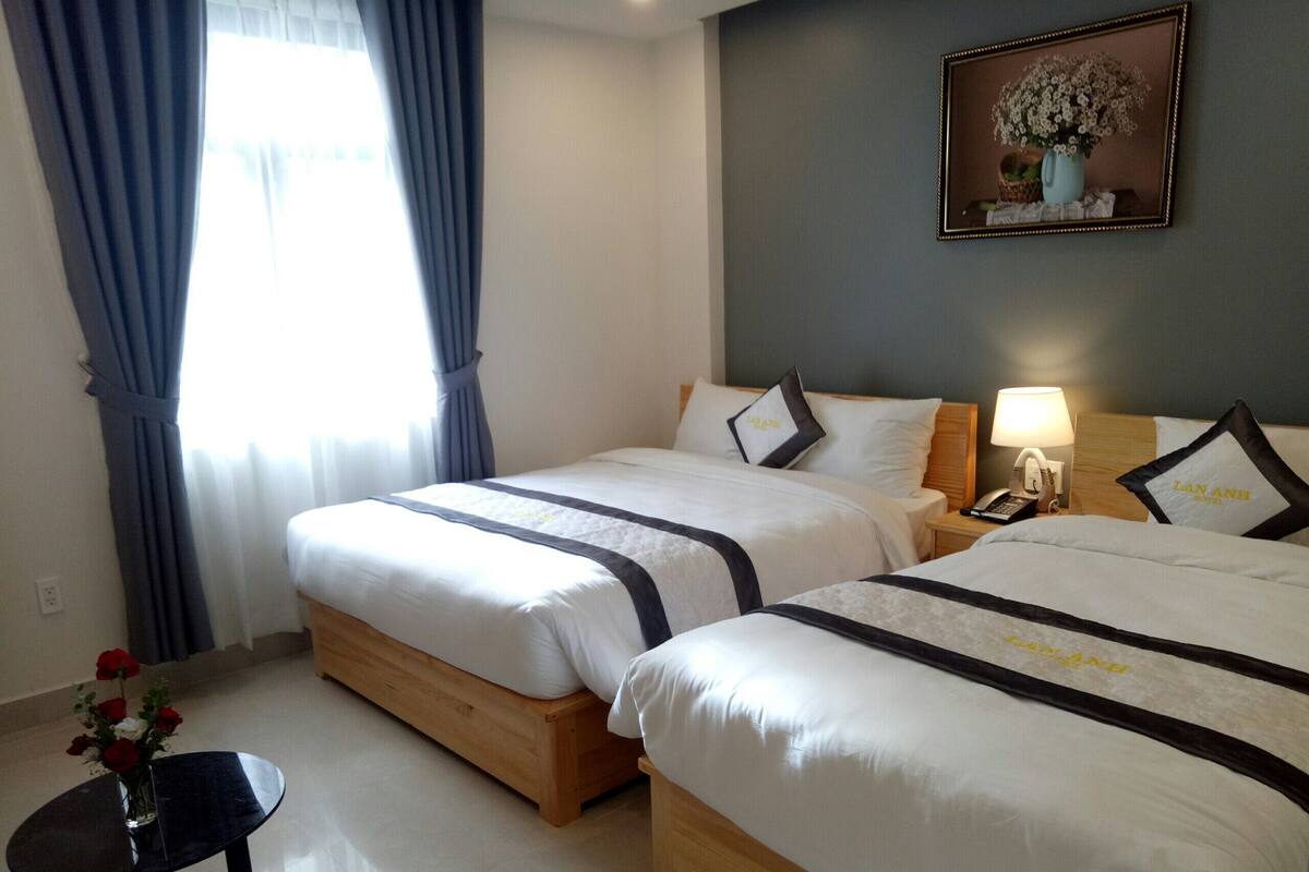 A hotel room with two beds, a bedside table with a lamp, a small table with flowers, and a framed picture on the wall.