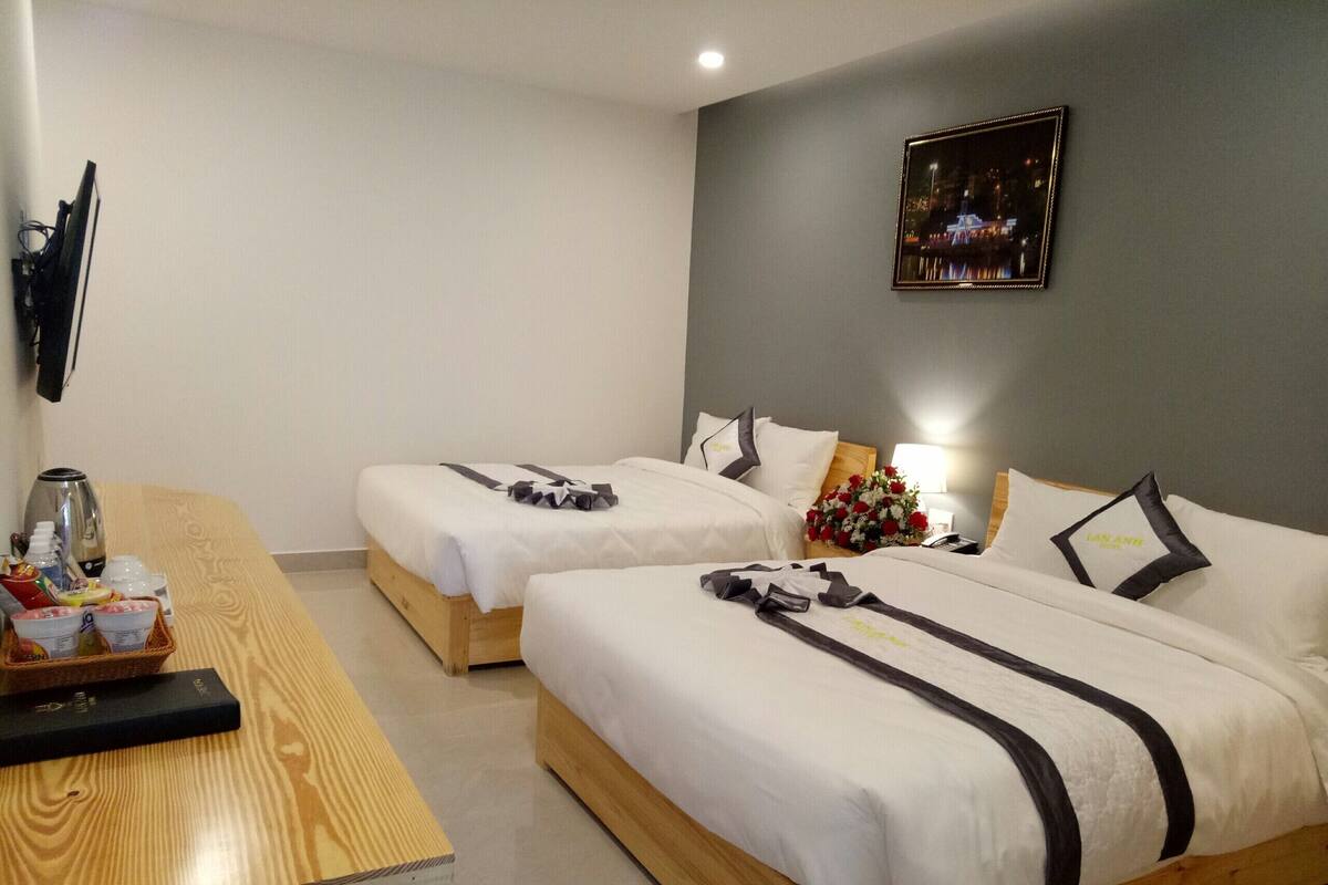 A hotel room with two beds, a wooden table with breakfast items, a nightstand with a lamp and a vase of flowers, and a framed picture on the wall.