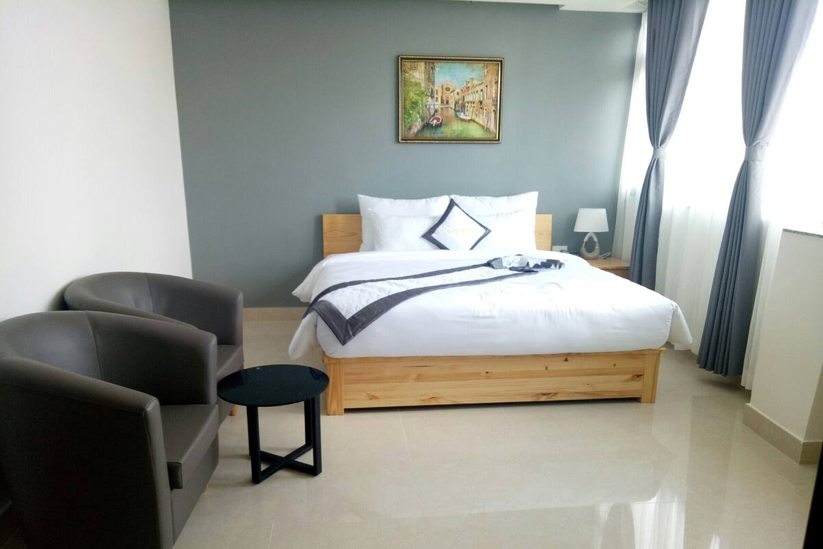 A hotel room with a bed, a chair, a small table, and a painting on the wall.