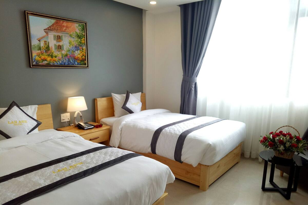 A hotel room with two beds, a nightstand with a lamp, a painting on the wall, and a window with curtains.