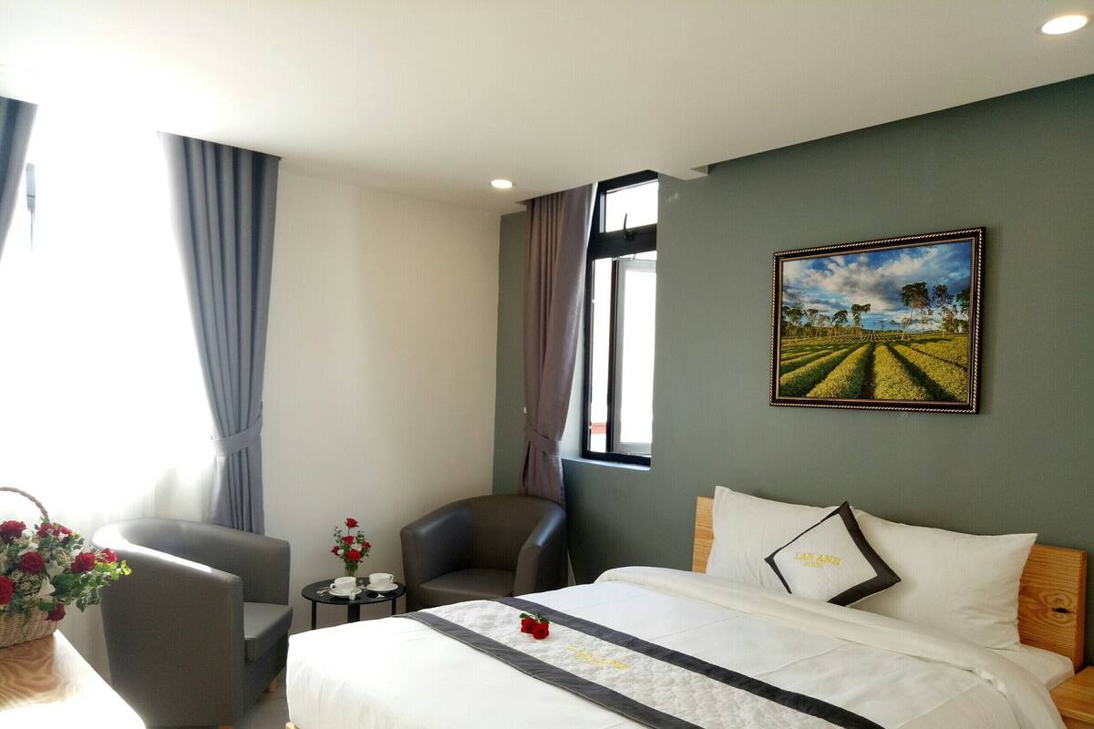 A hotel room with a bed, a chair, a small table with a vase of flowers, and a painting on the wall.