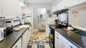 Eine kompakte Küche mit weißen Schränken, einer Mikrowelle, einem Herd und einem Spülbecken. Ein Teppich mit dem Aufdruck 'GETAWAY' und eine kleine Sitzecke mit einer Bank und dekorativen Elementen.