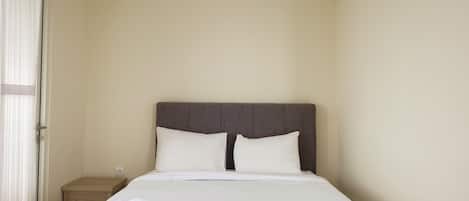 A neatly made bed with white linens and two pillows, a wooden nightstand with a lamp, and a door with a curtain.