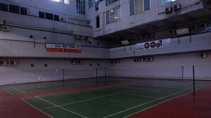 Ein Hallen-Tennisplatz mit grüner und roter Oberfläche, umgeben von Wänden mit Klimaanlagen und einem Schild, das ein Hotel bewirbt.