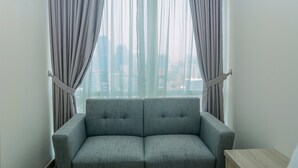 A room with a grey sofa, a wooden side table, and a window with curtains. - Modern and Cozy Studio Menteng Park Apartment (Jakarta)