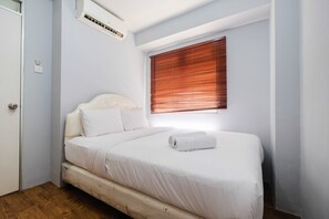 Une chambre avec un lit simple, un plancher en bois, du linge de lit blanc, une fenêtre avec des stores et un appareil de climatisation.