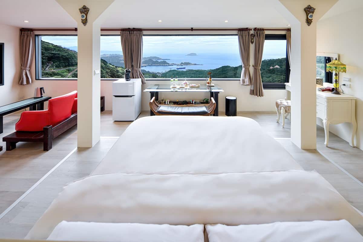 A spacious bedroom with a large bed, a desk with a chair, a small table, and a view of a mountainous landscape.