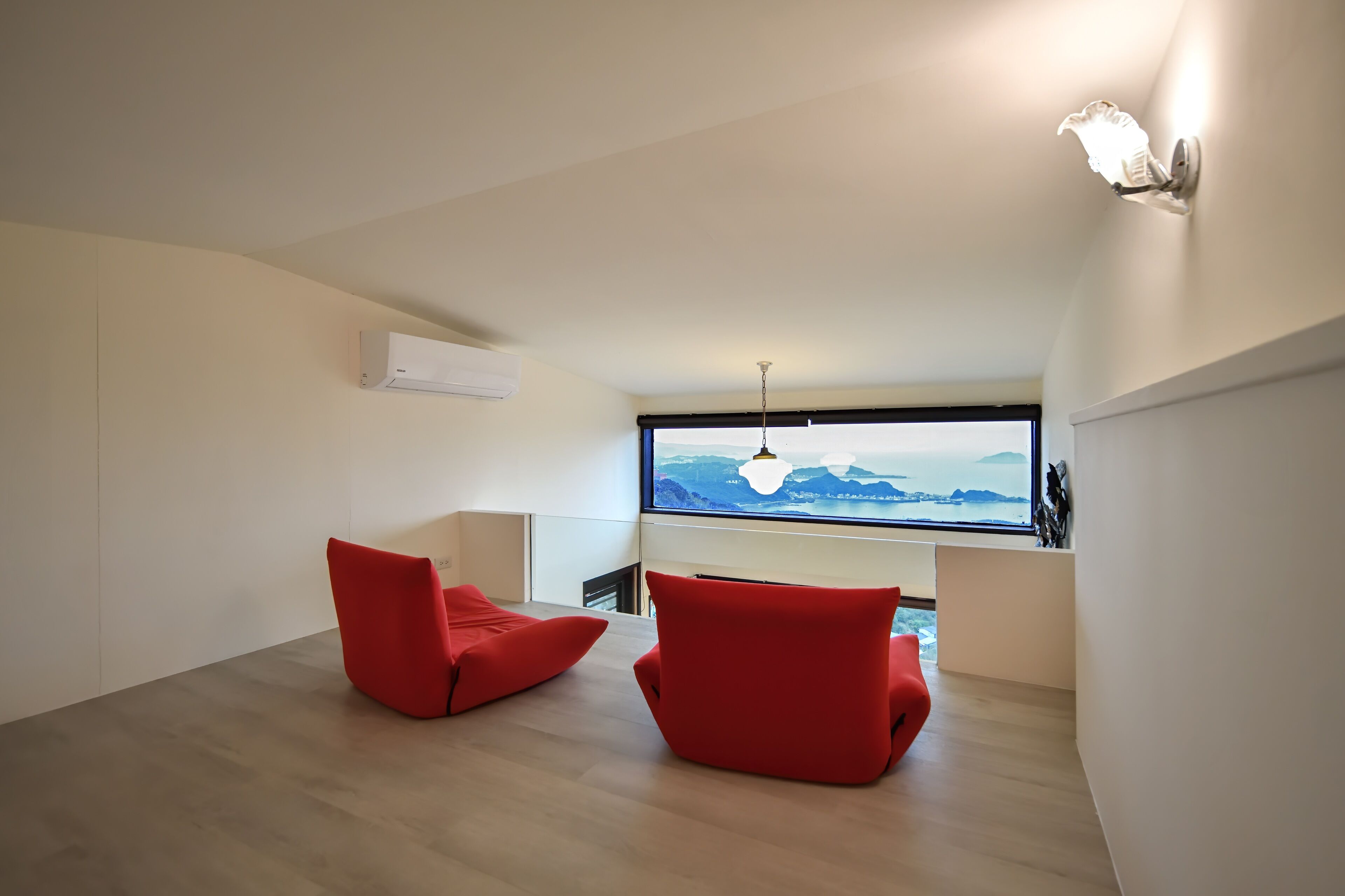 A modern room with a large window, two red chairs, a hanging light fixture, and a white air conditioning unit.