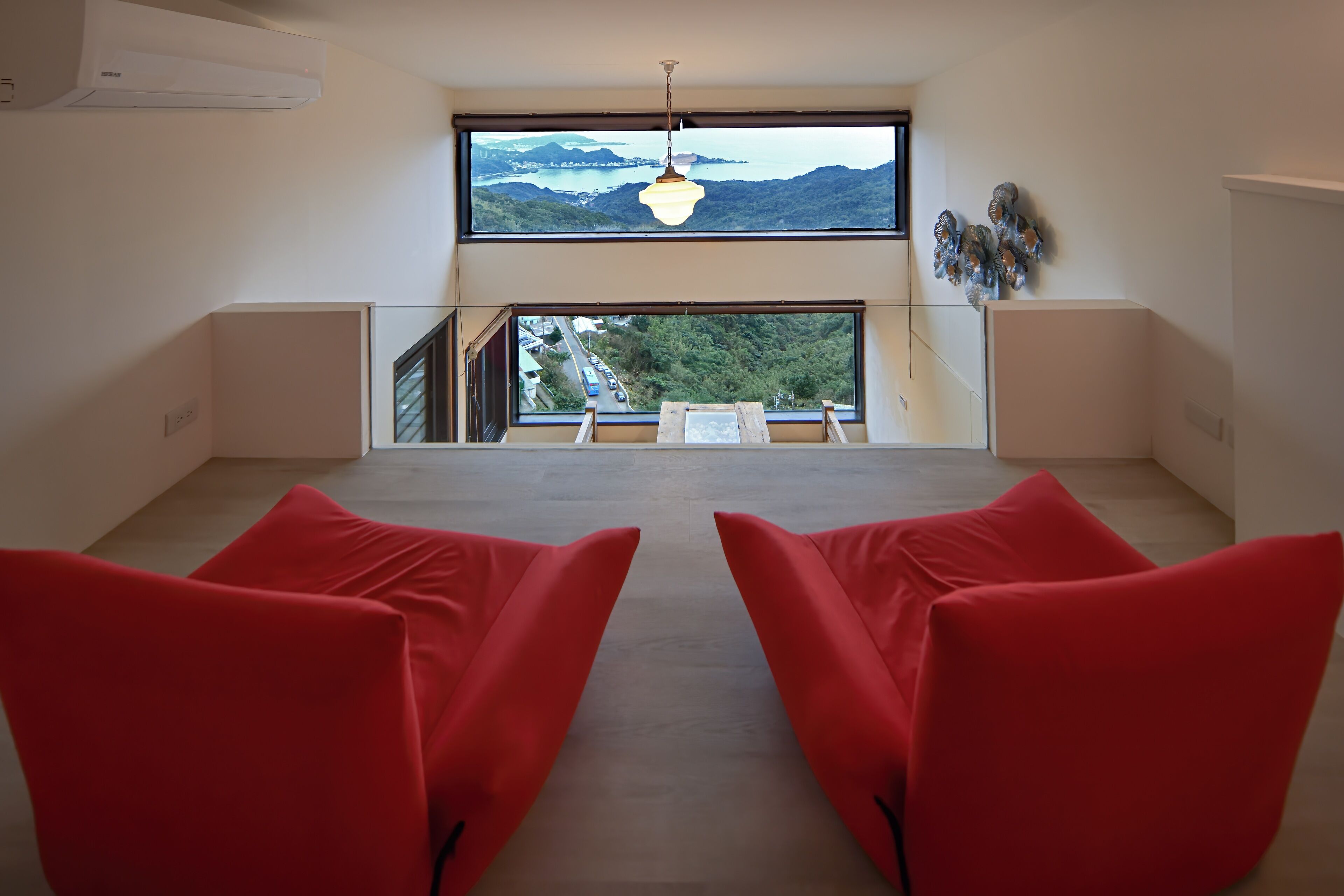 A room with a large window offering a view of mountains and a body of water. There are two red chairs and a hanging light fixture.