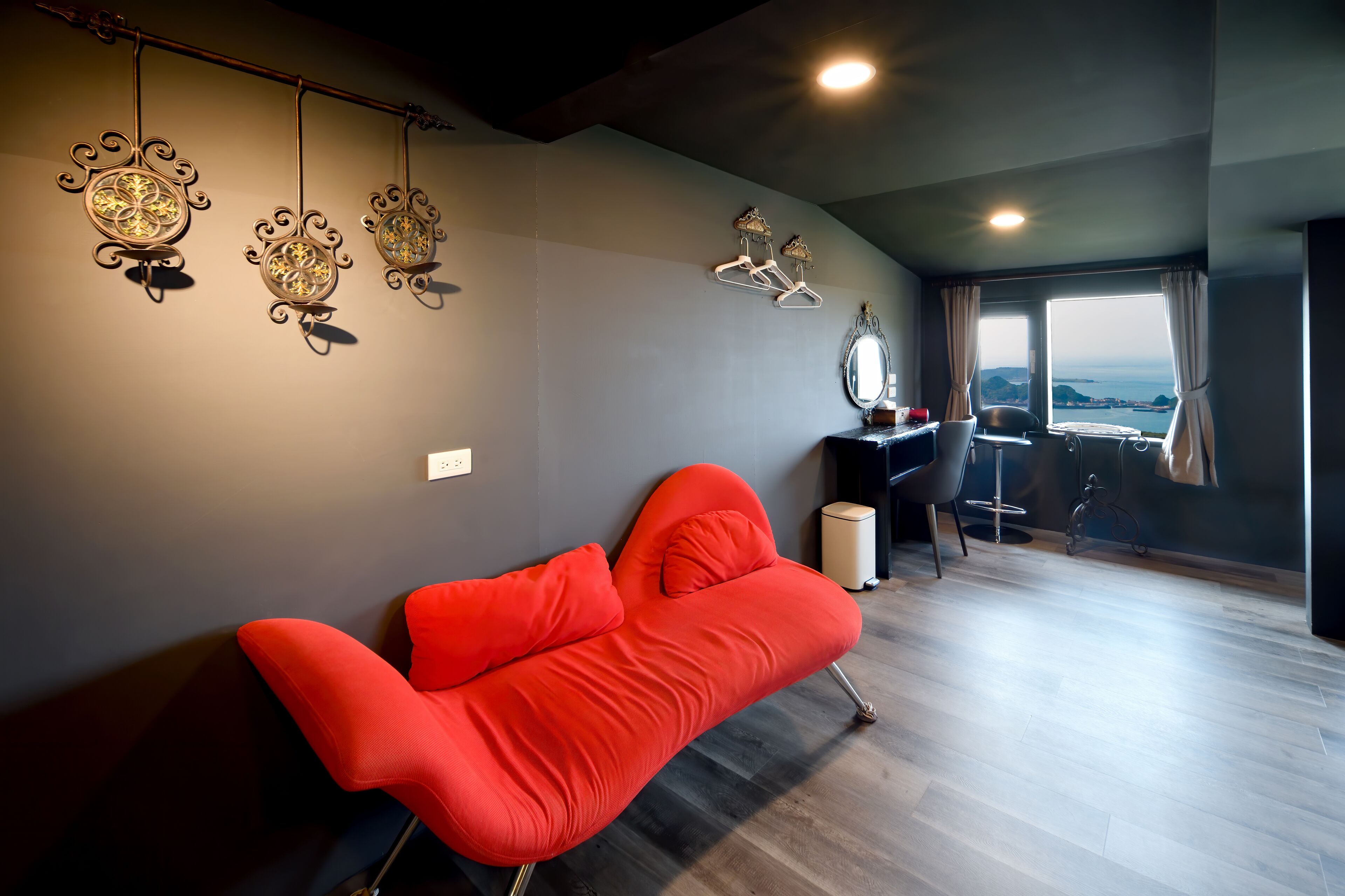 A modern living room with a red sofa, decorative wall hangings, a desk with a chair, and a view of the sea.