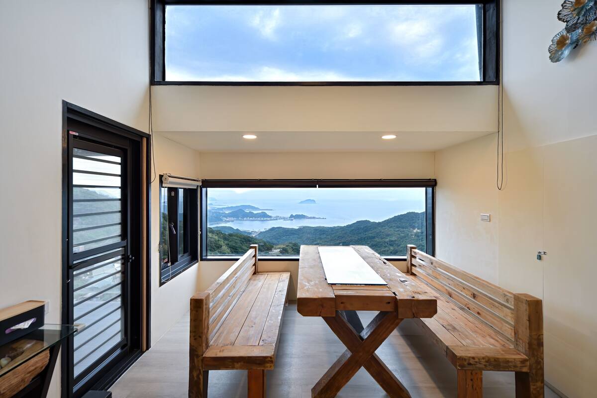 A modern room with a large window offering a view of hills and a body of water. There are two wooden benches and a table in the center.