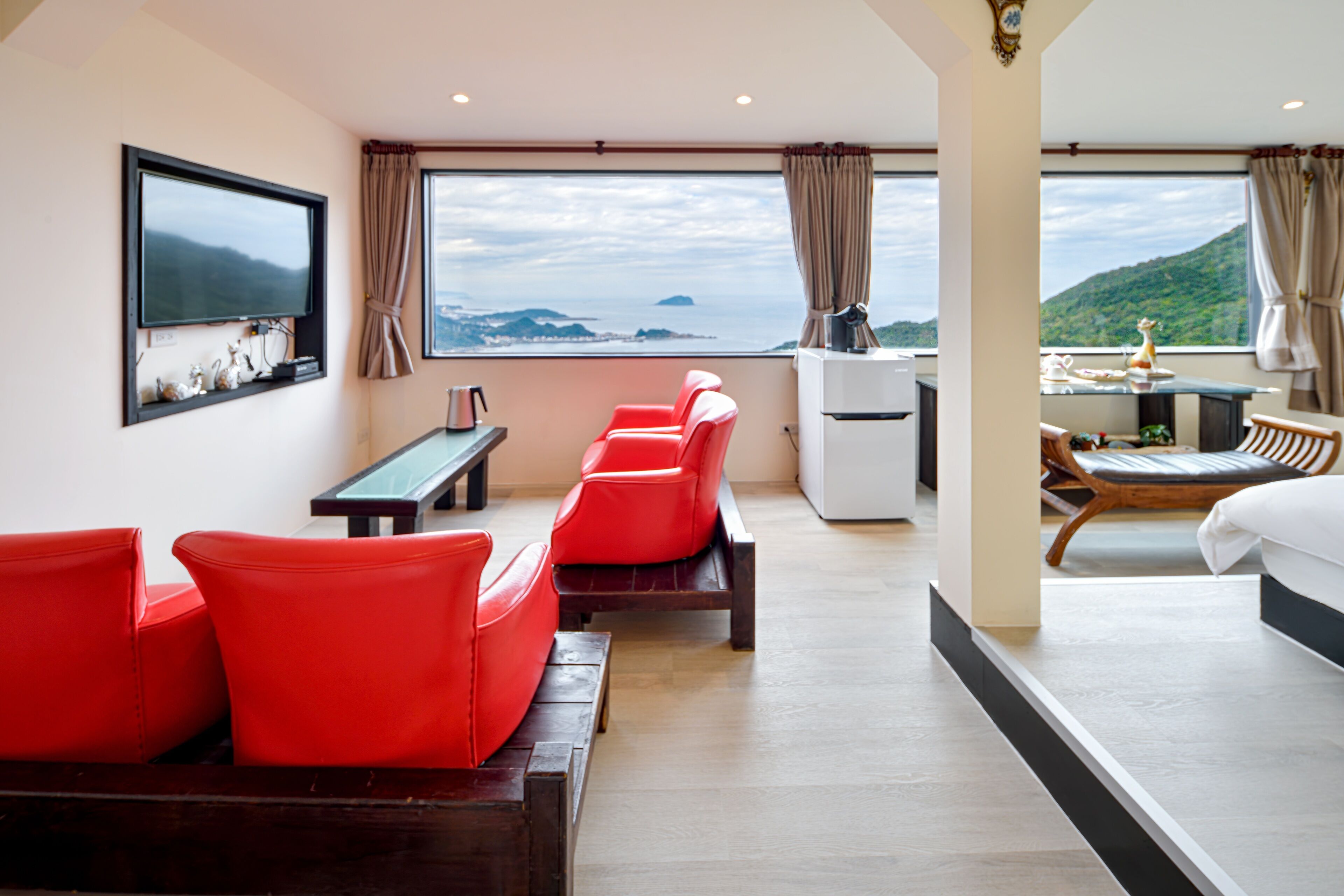 A modern hotel room with a large window, red seating, a flat-screen TV, and a dining area with a view.