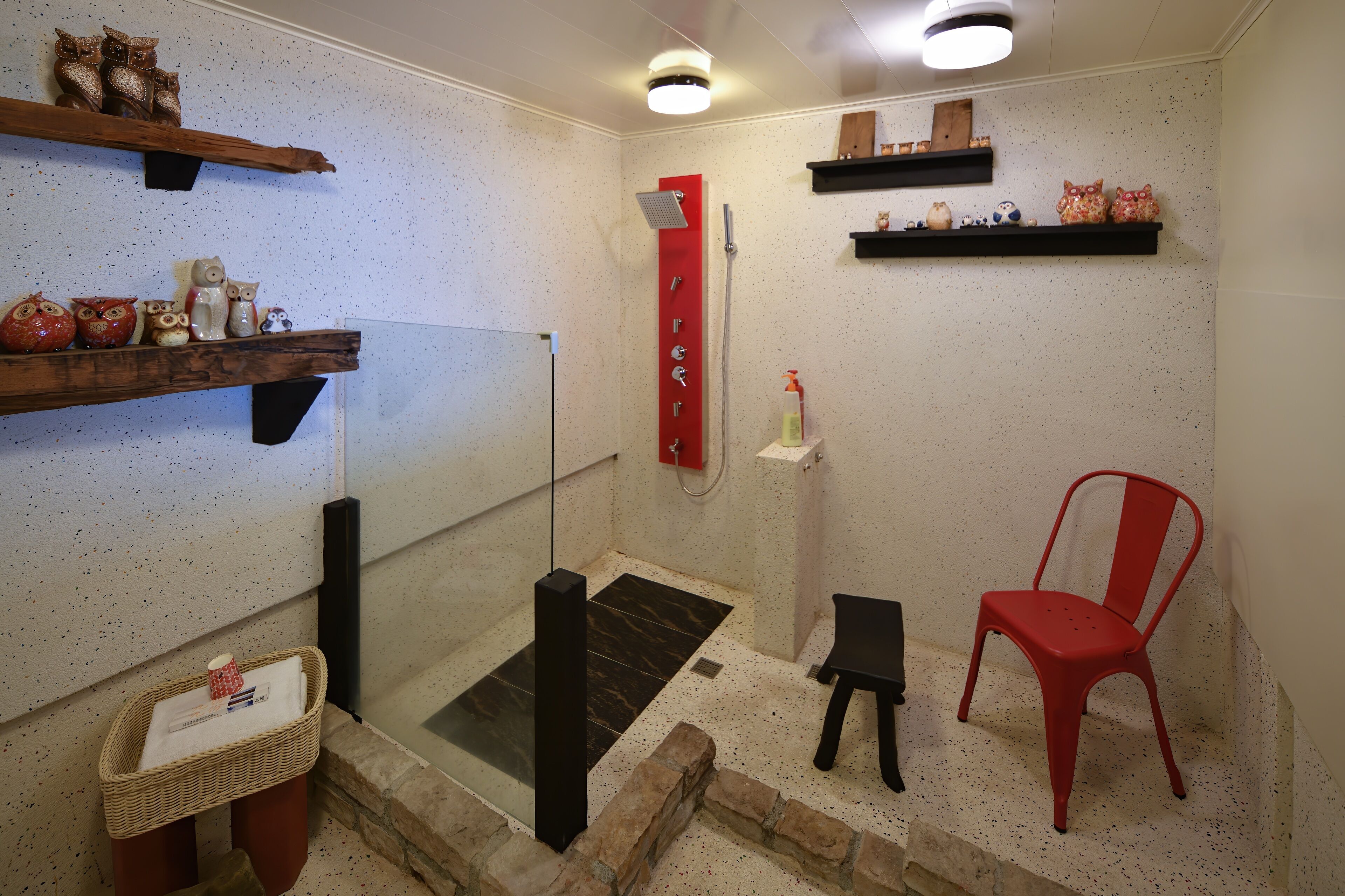 A shower area with a glass door, a red chair, and shelves with decorative items.