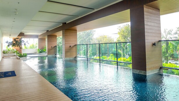 An indoor swimming pool with clear glass walls, wooden pillars, and a view of greenery outside. - Spacious Studio Room at Capitol Suites Apartment (Jakarta)