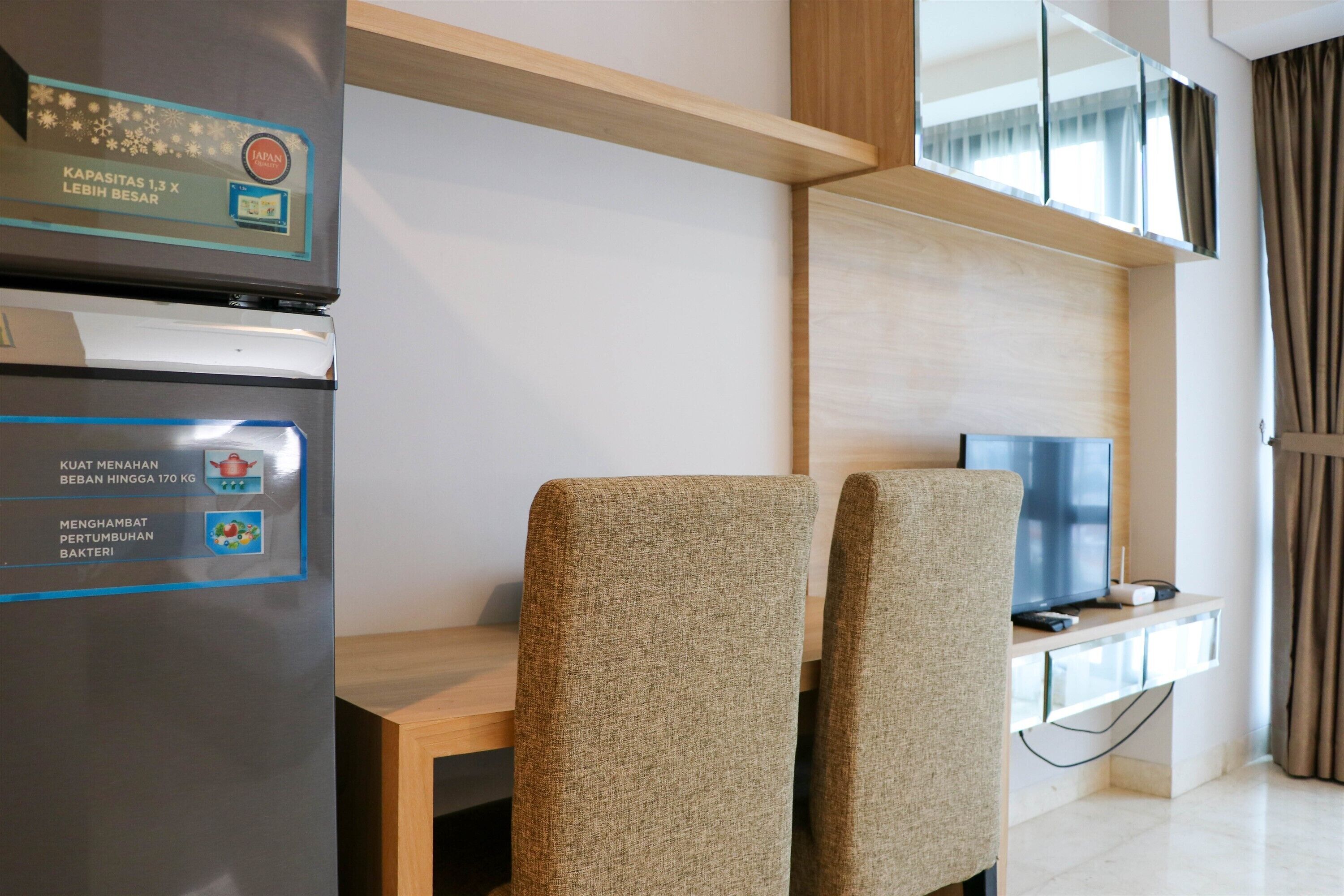 Una habitación de hotel moderna con un refrigerador incorporado, un televisor de pantalla plana sobre un soporte de madera y dos sillones tapizados.