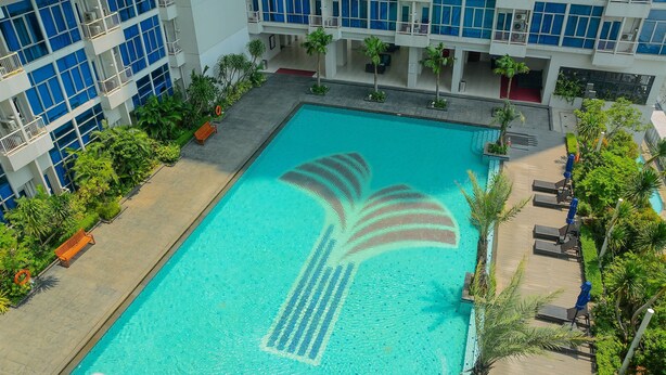 Una piscina con diseño de palmera, rodeada de sillones de descanso y palmeras, con un edificio al fondo.