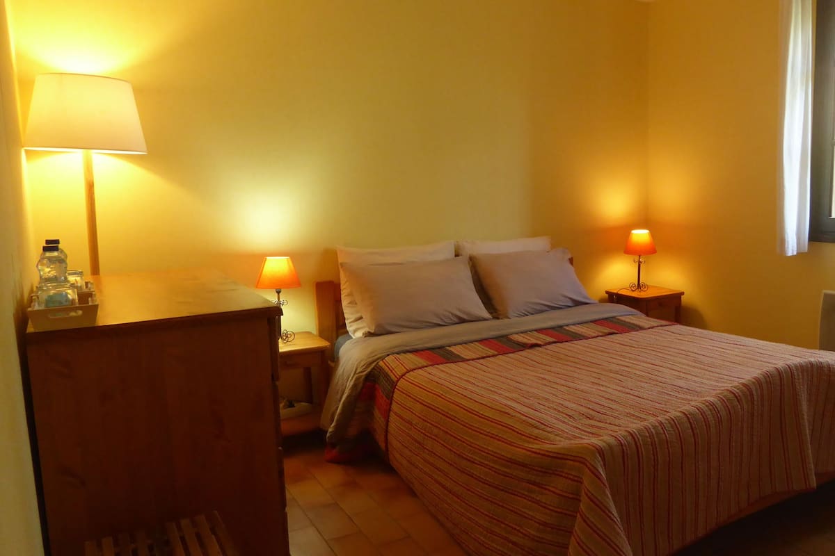 A bedroom with a bed, two bedside tables with lamps, a wooden nightstand, and a window with blinds.