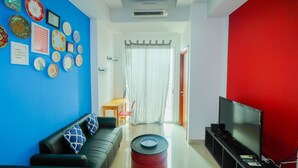 A living room with a blue accent wall, a black leather sofa, a red barrel, a flat-screen TV, and a dining area with a table and chairs. - Minimalist 2BR Apartment at Springhill Terrace Residence (Jakarta)