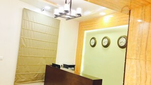A reception area with a wooden reception desk, a large window with blinds, and two wall clocks. - Hotel Ajmer Regency (Ajmer)