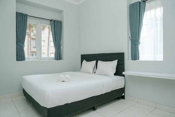 A neatly made bed with white linens and pillows, a dark headboard, and a window with curtains.