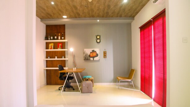 A modern room with a wooden ceiling, a desk with a chair, a small table, a bench, a red curtain, and a shelf with bottles and a clock.
