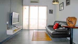 A modern living room with a flat-screen TV, a leather sofa, a wooden side table, and a large window with blinds.