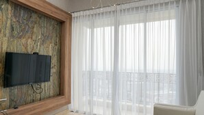 Un salón moderno con un televisor de pantalla plana montado en una pared con textura, un sofá blanco y un balcón con cortinas traslúcidas.