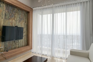 Un salón moderno con un televisor de pantalla plana montado en una pared con textura, un sofá blanco y un balcón con cortinas traslúcidas.