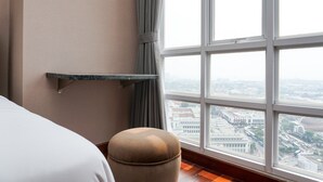 Habitación de hotel con cama, un banco acolchado, una ventana con vista a la ciudad y un estante en la pared.