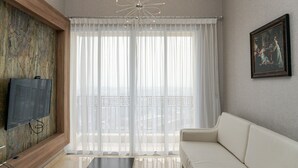 Una sala de estar moderna con un televisor de pantalla plana montado en una pared texturizada, un sofá blanco, una mesa de centro y un amplio ventanal con cortinas translúcidas.