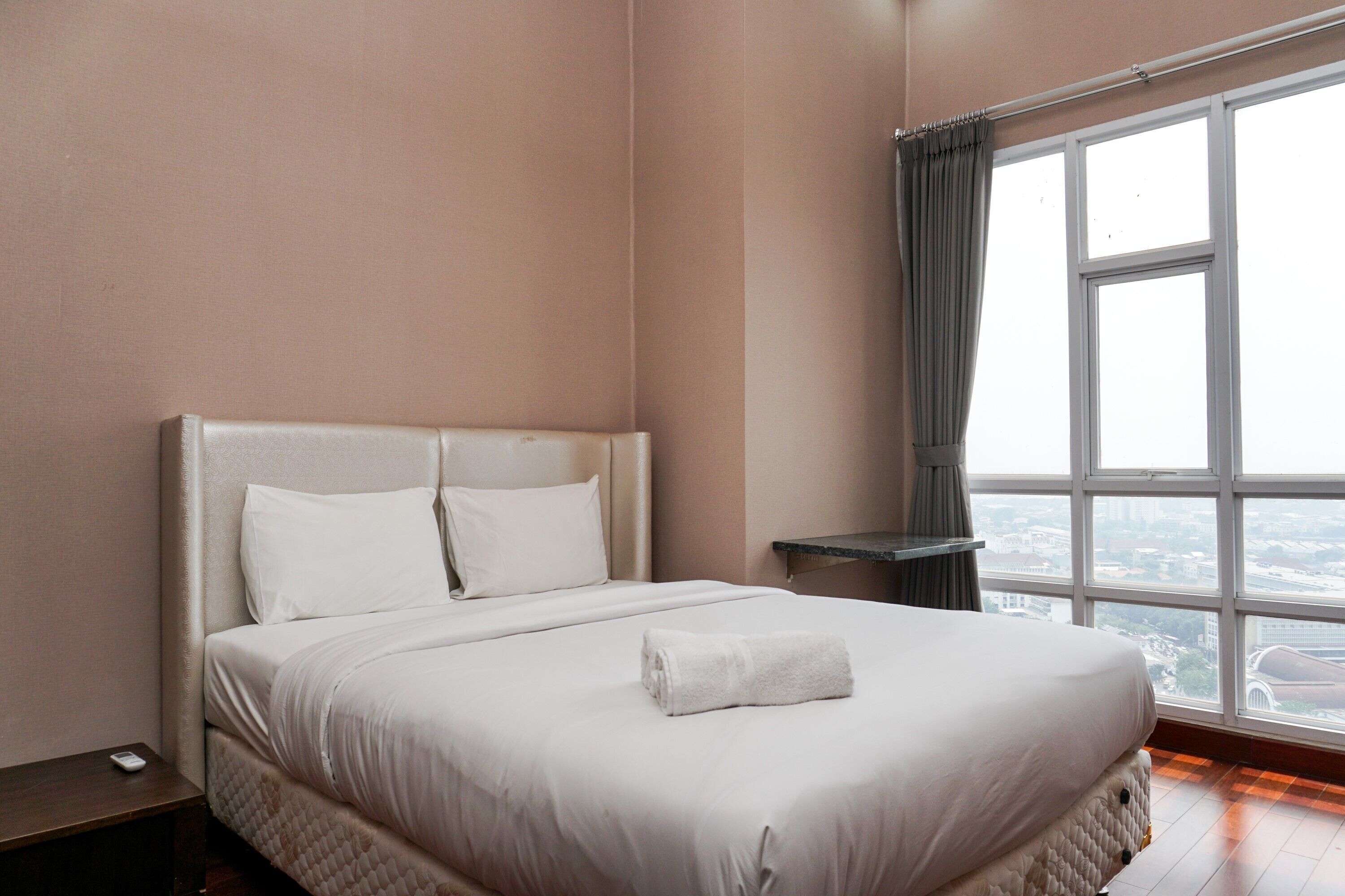 A neatly made bed with white linens and a rolled towel, a bedside table with a remote control, and a large window with a view of the city.