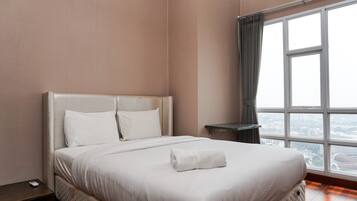 A neatly made bed with white linens and a rolled towel, a bedside table with a remote control, and a large window with a view of the city.