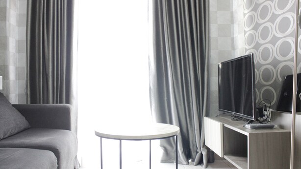A modern living room with a grey sofa, a small round table, a TV on a wooden stand, and grey curtains. - Modern 2BR Apartment at Mekarwangi Square Cibaduyut (Bandung)