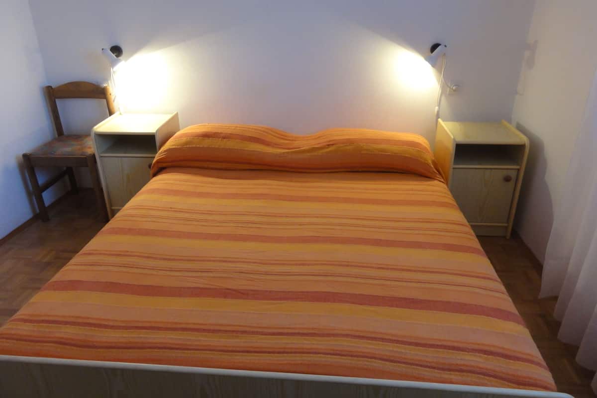Uma cama com colcha listrada, duas luminárias de cabeceira, uma cadeira de madeira e uma mesa de cabeceira com uma gaveta.