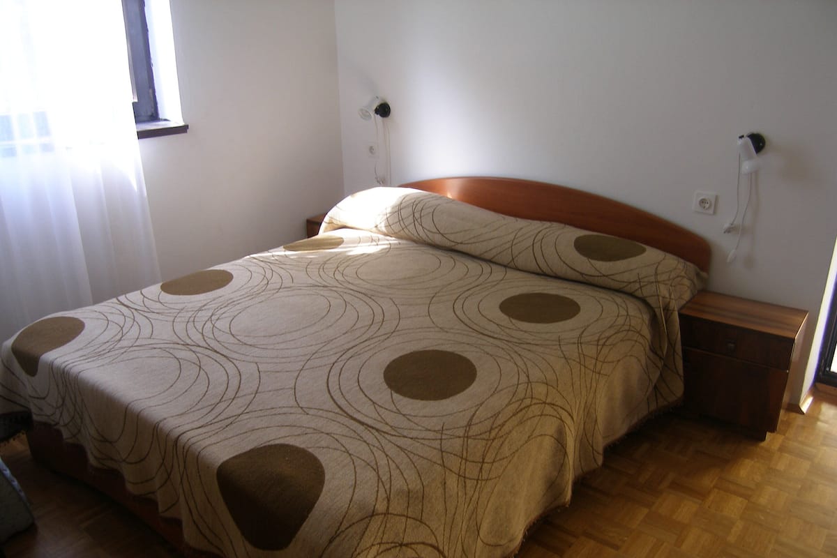 Quarto com cabeceira de madeira, cama com colcha estampada, mesa de cabeceira e uma tomada na parede.