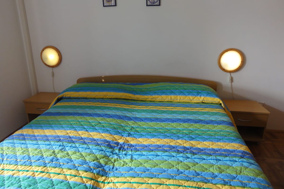 Uma cama com um colcha colorida, duas mesas de cabeceira com abajures e duas fotos emolduradas na parede.