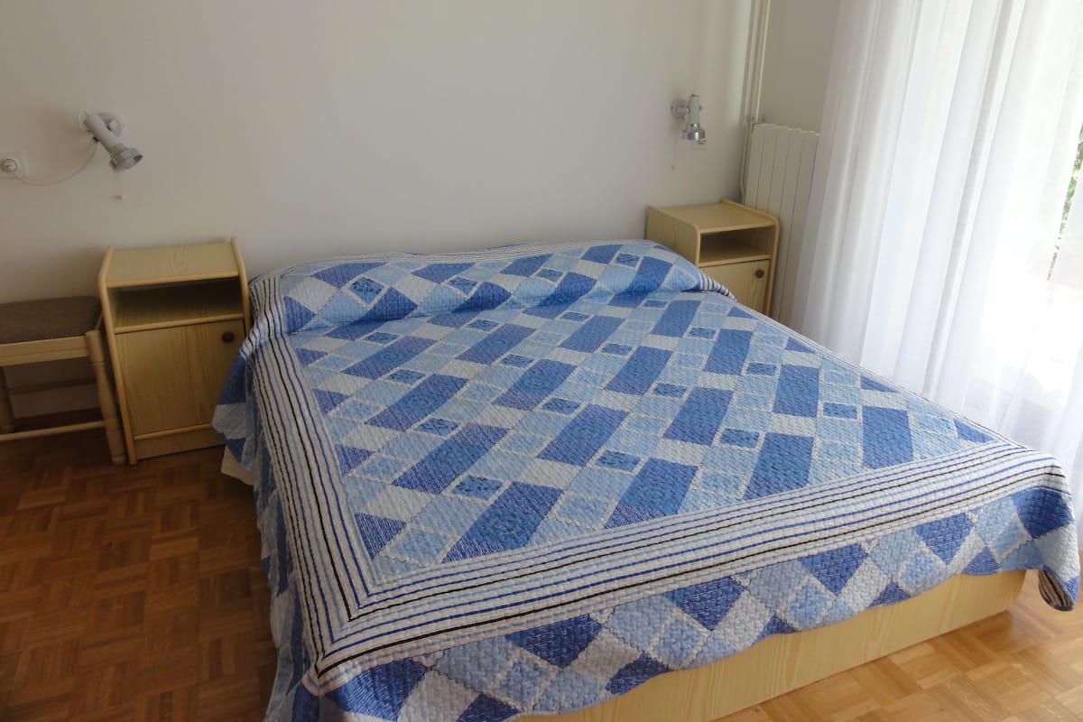 Quarto com cama de madeira, duas mesas de cabeceira, janela com cortinas finas e radiador.