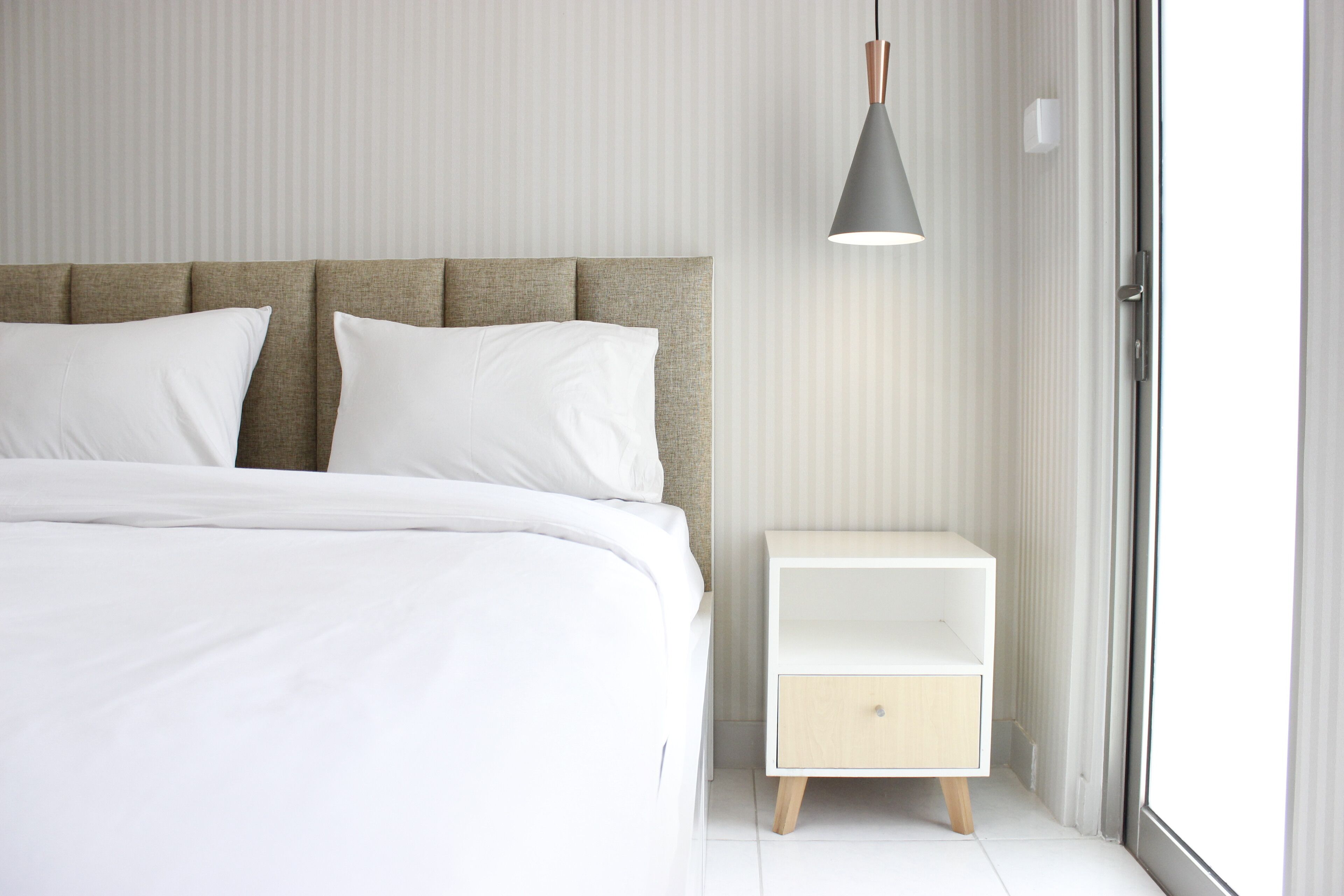 A neatly made bed with white linens and a headboard, a bedside table with a drawer, and a hanging light fixture.