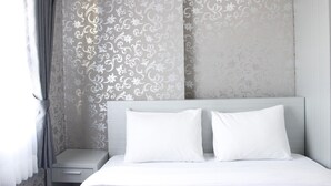 A neatly made bed with white bedding and pillows, a bedside table, and a wall with a floral pattern. - Stylish 2BR Gateway Pasteur Apartment (Cimahi)