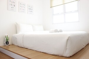 Camera da letto minimalista con un letto grande, biancheria bianca, una piccola decorazione di cactus e un'opera d'arte a parete raffigurante diverse piante.