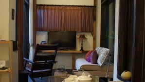A living room with a sofa, a coffee table, and a TV. There is a window with curtains, a lamp, and a small shelf with items.