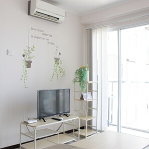 A modern living room with a flat-screen TV on a white stand, a small table with a remote control, and a shelving unit with a potted plant.