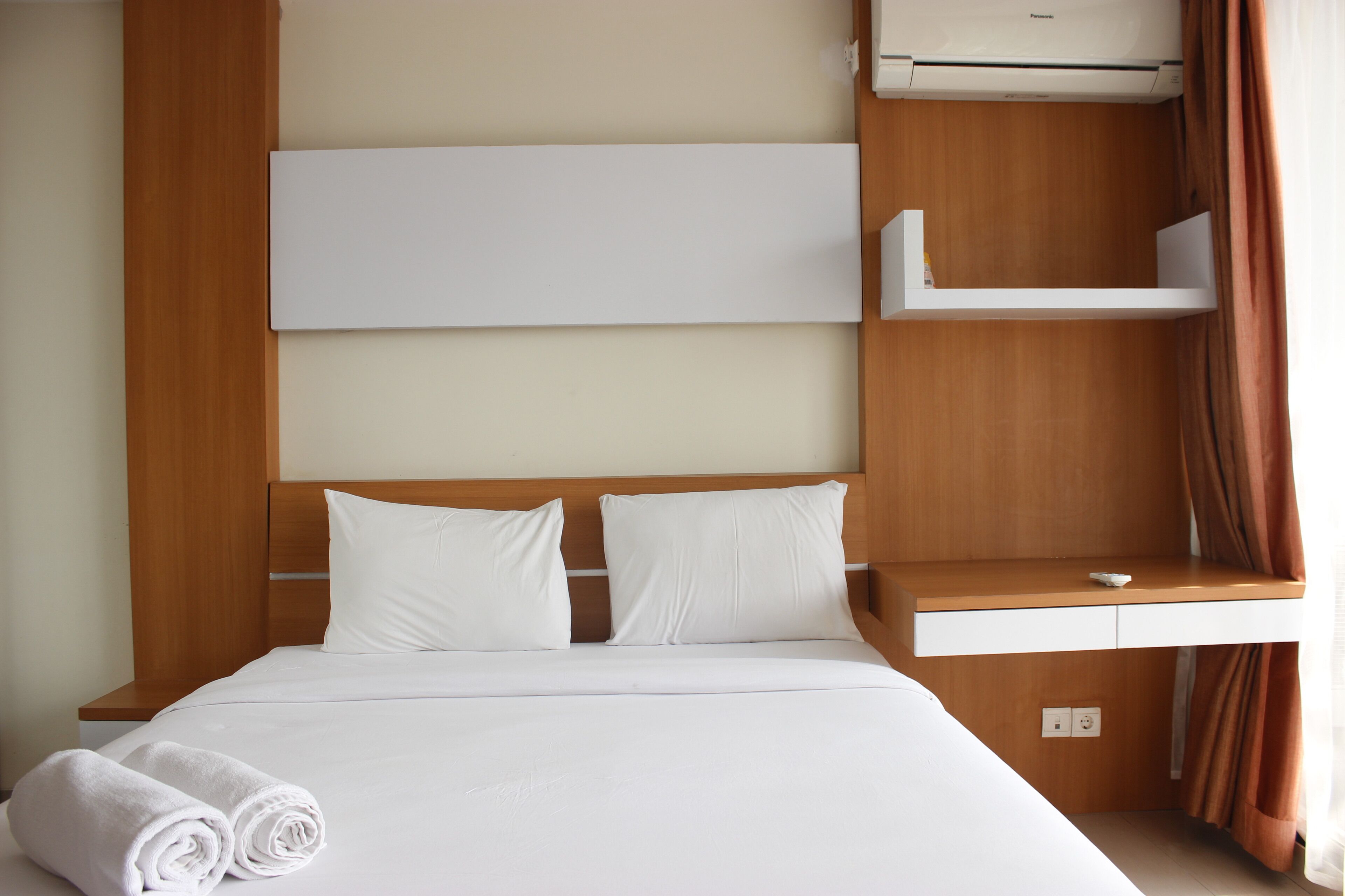A hotel room with a wooden headboard, white bedding, a small white shelf, and an air conditioning unit.