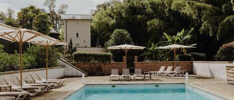 Une piscine entourée de chaises longues et de parasols, avec de la verdure tout autour et un bâtiment en arrière-plan.