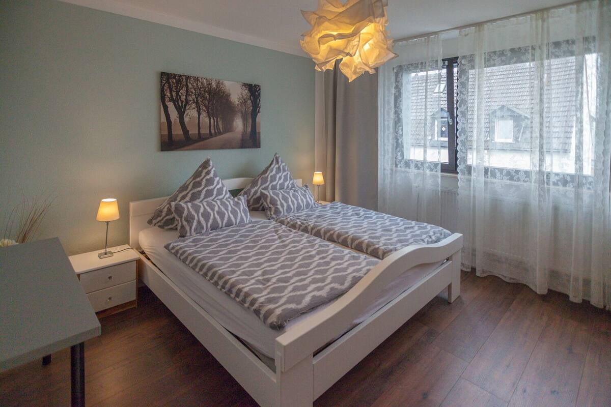 Ein Schlafzimmer mit Bett, Nachttischen, einer Lampe, einem Gemälde und einem Fenster mit Vorhängen.