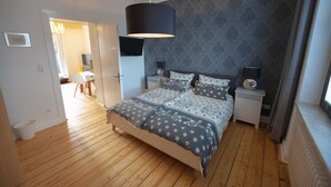 A bedroom with a wooden floor, a bed with patterned bedding, a wall-mounted TV, bedside lamps, and a window with curtains. - Apartmenthaus Lorbach (Solingen)