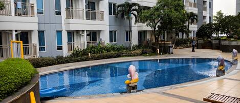 A swimming pool with a statue, surrounded by a building with balconies and a tiled walkway.