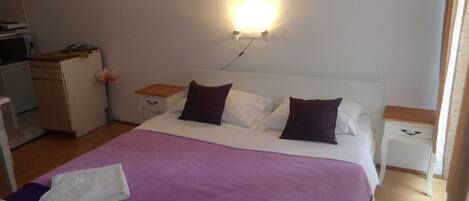 A neatly made bed with a purple bedspread and white towels, a bedside table with a lamp, and a small wooden cabinet.