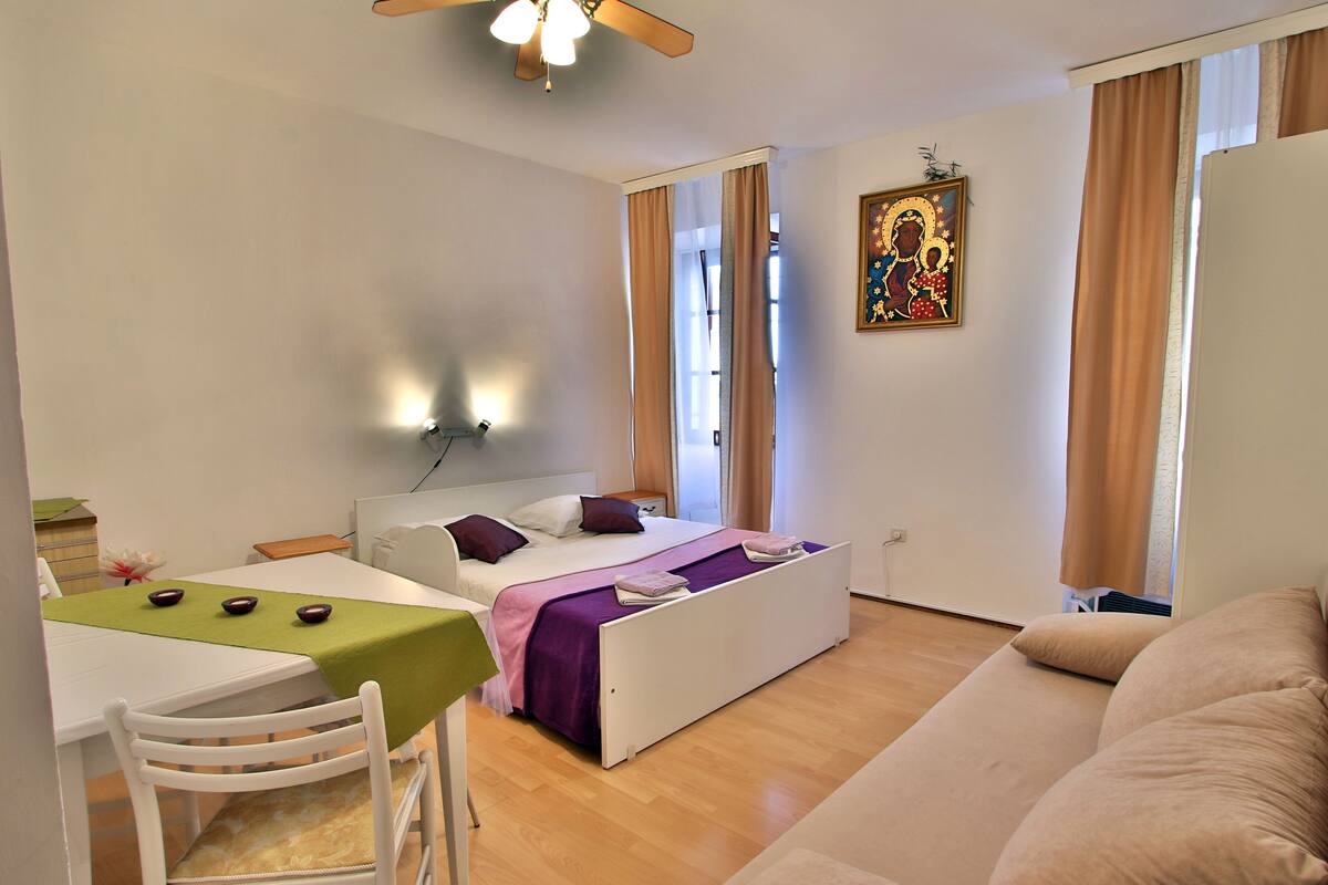 Un dormitorio con cama, escritorio y silla. Hay una ventana con cortinas, un ventilador de techo y una lámpara de pared.