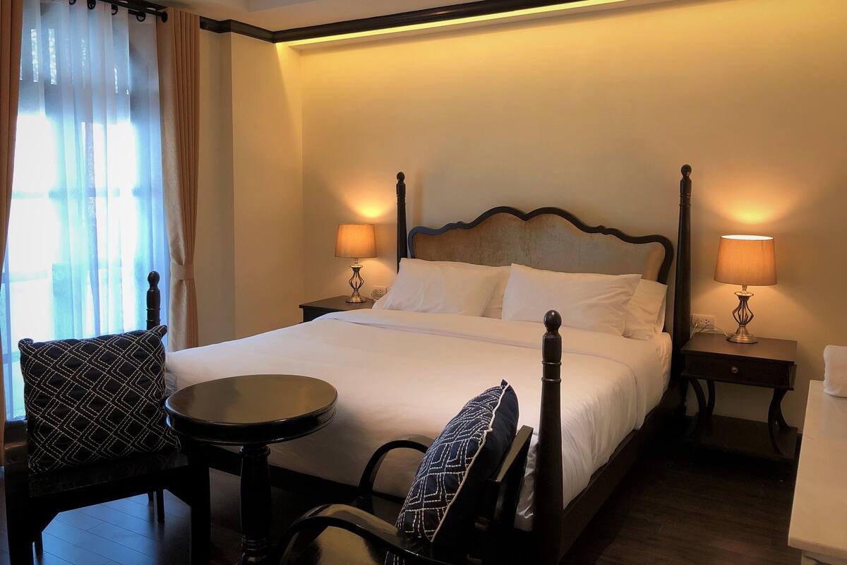 Une chambre d’hôtel bien aménagée, avec un lit à baldaquin, deux tables de chevet avec des lampes, une table ronde et deux chaises.